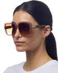 A woman with straight, dark hair tied back wears Okkia Oversize-Stil Sonnenbrille with UV 400-Schutz, featuring a pinkish-brown translucent frame and tortoiseshell-patterned arms. She has a neutral expression and wears a plain white top against a white background.