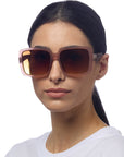 A woman with straight dark hair pulled back wears Okkia Oversize-Stil Sonnenbrille with rose-tinted, brown gradient lenses and UV 400-Schutz. She has a neutral expression and a white shirt against a plain white background.