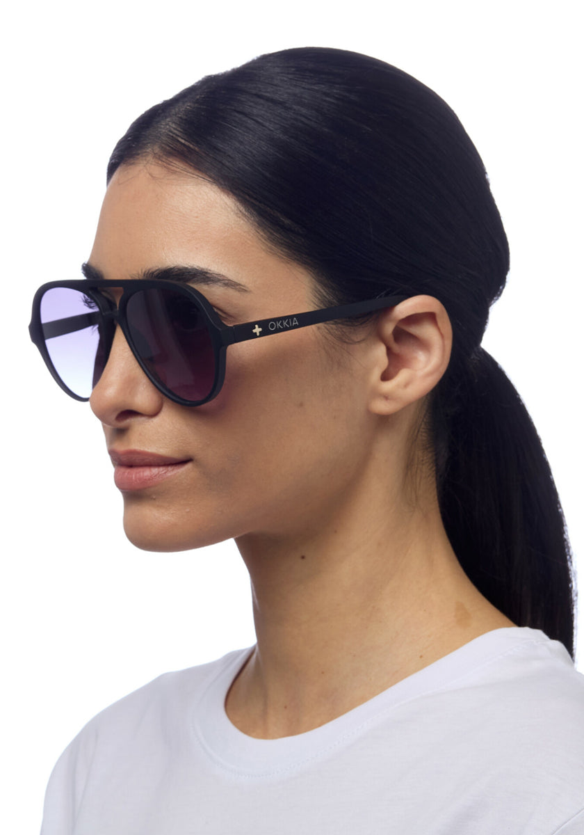 A woman with straight black hair in a low ponytail wears oversized, black-rimmed Okkia sunglasses featuring UV 400-Schutz and gold brand lettering on the side. She is shown in profile against a white background, dressed in a white shirt.