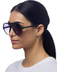 A woman with straight black hair in a low ponytail wears oversized, black-rimmed Okkia sunglasses featuring UV 400-Schutz and gold brand lettering on the side. She is shown in profile against a white background, dressed in a white shirt.