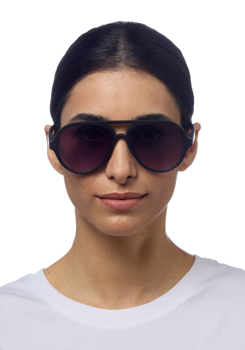 A woman with dark hair pulled back wears large, round Okkia black sunglasses with UV 400 protection and a white crew-neck shirt, facing forward and smiling slightly against a plain white background.
