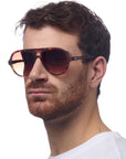 A man with short brown hair and a trimmed beard wears large, brown tortoiseshell aviator sunglasses by Okkia. He faces left in a white shirt with a neutral expression against a plain white background.