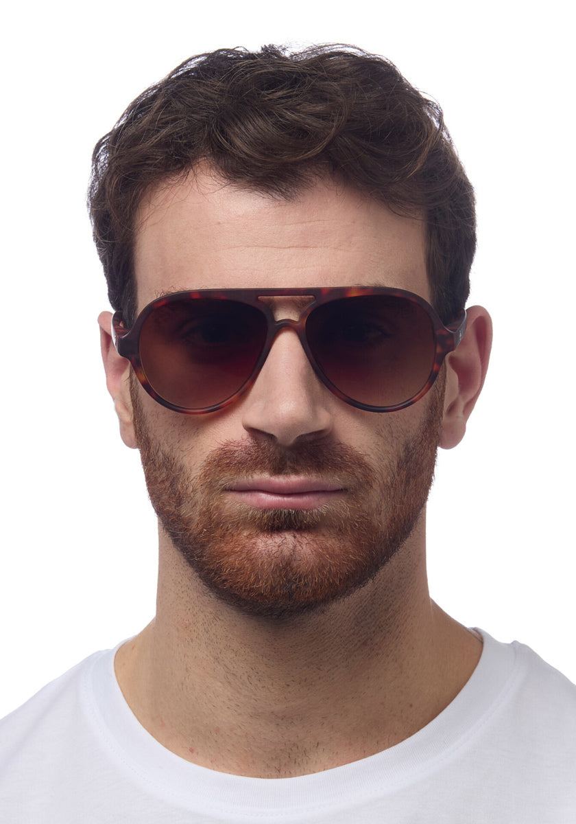 A man with short brown hair and a trimmed reddish-brown beard wears large, round Okkia Aviator-style sunglasses with UV 400 protection and a white crew-neck shirt, facing forward against a plain white background.
