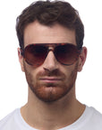 A man with short brown hair and a trimmed reddish-brown beard wears large, round Okkia Aviator-style sunglasses with UV 400 protection and a white crew-neck shirt, facing forward against a plain white background.