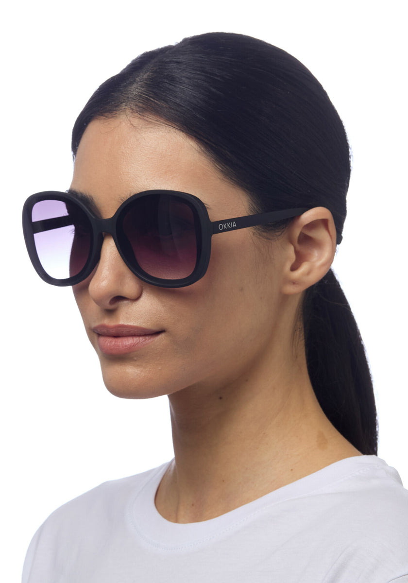 A woman with straight dark hair tied back wears oversized Okkia ANNA butterfly sunglasses with gradient lenses and UV protection, featuring the Okkia logo on the side. She pairs them with a white top against a plain white background.