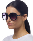 A woman with straight dark hair tied back wears oversized Okkia ANNA butterfly sunglasses with gradient lenses and UV protection, featuring the Okkia logo on the side. She pairs them with a white top against a plain white background.