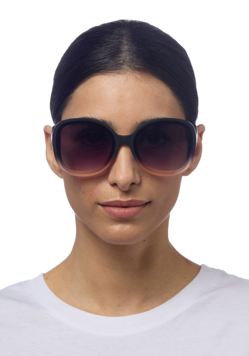 A woman with dark hair pulled back wears oversized black Okkia ANNA sunglasses in butterfly style and a white crew-neck shirt, facing forward with a neutral expression against a plain white background.
