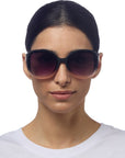 A woman with dark hair pulled back wears oversized black Okkia ANNA sunglasses in butterfly style and a white crew-neck shirt, facing forward with a neutral expression against a plain white background.