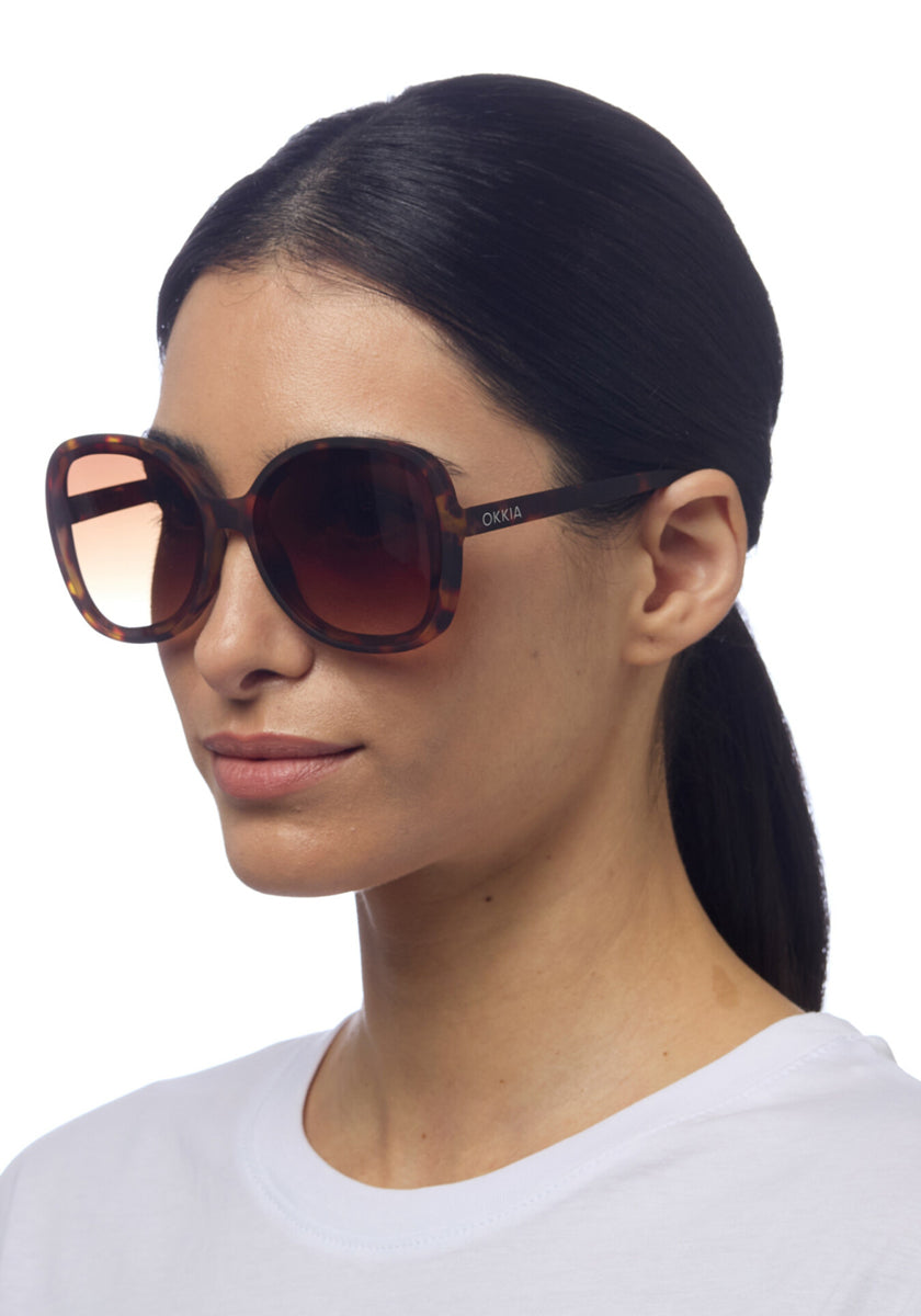 A woman with straight dark hair pulled back wears oversized, round Okkia ANNA tortoiseshell sunglasses with UV protection and a plain white shirt. Posed against a white background, she faces slightly left, revealing OKKIA on the sunglass arm.