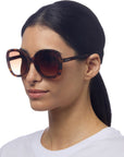 A woman with straight dark hair pulled back wears oversized, round Okkia ANNA tortoiseshell sunglasses with UV protection and a plain white shirt. Posed against a white background, she faces slightly left, revealing OKKIA on the sunglass arm.