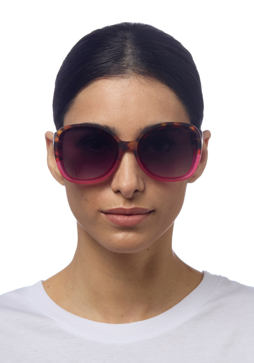A woman with dark hair pulled back wears oversized, round Okkia ANNA sunglasses in butterfly style with a pink and tortoiseshell frame. She has a neutral expression, a plain white shirt, a white background, and enjoys included UV protection.