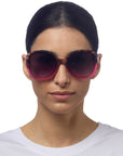 A woman with dark hair pulled back wears oversized, round Okkia ANNA sunglasses in butterfly style with a pink and tortoiseshell frame. She has a neutral expression, a plain white shirt, a white background, and enjoys included UV protection.