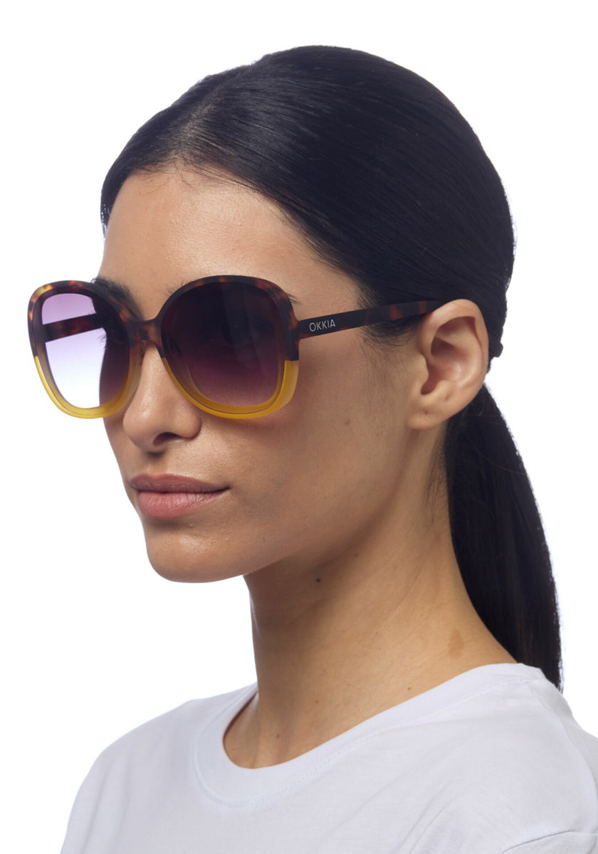 A woman with straight, dark hair tied back wears large, round Okkia Butterfly-Design Sonnenbrille with getönten Verlaufsgläsern and brown-yellow gradient frames. She has a neutral expression, a plain white top, and stands against a white background.
