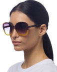 A woman with straight, dark hair tied back wears large, round Okkia Butterfly-Design Sonnenbrille with getönten Verlaufsgläsern and brown-yellow gradient frames. She has a neutral expression, a plain white top, and stands against a white background.