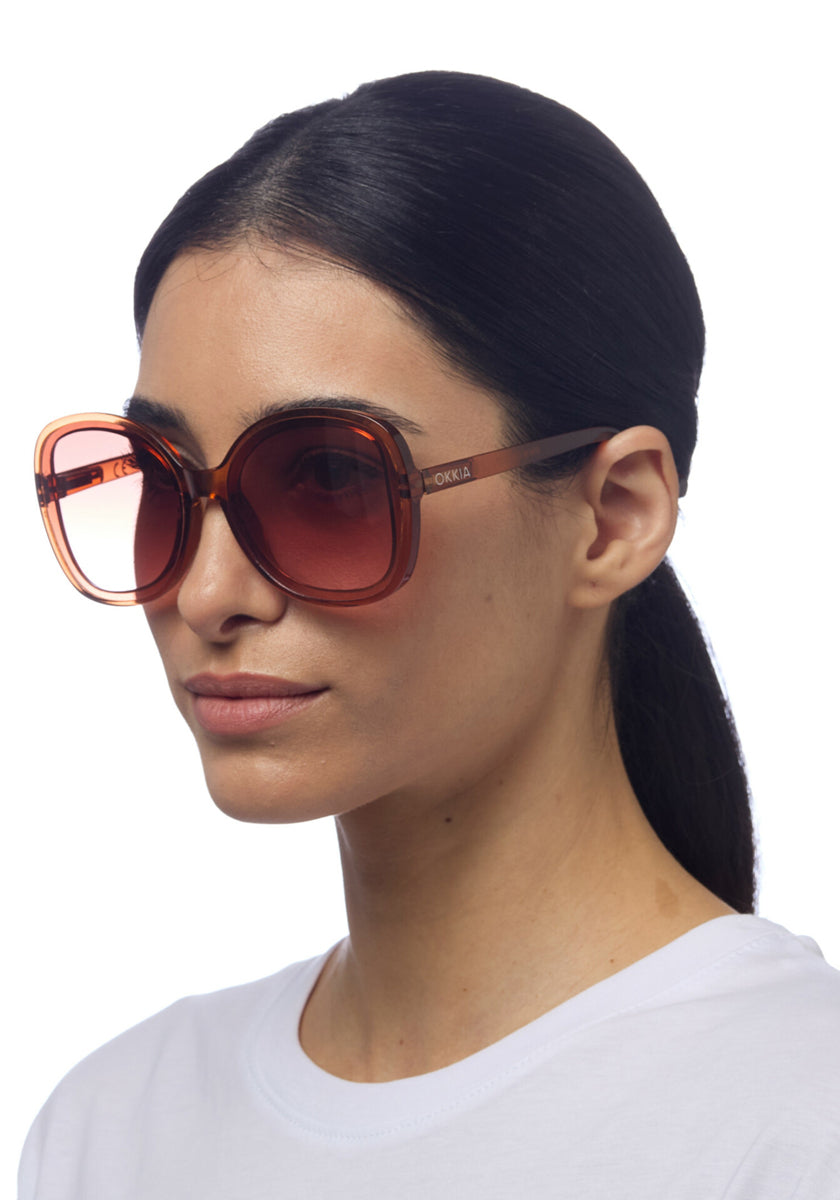 A woman with long dark hair tied back wears oversized Okkia Sonnenbrille im Butterfly-Stil with translucent amber frames and gradient lenses. She has a neutral expression, a plain white crew-neck shirt, and stands against a white background.