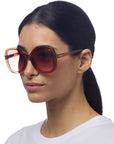 A woman with long dark hair tied back wears oversized Okkia Sonnenbrille im Butterfly-Stil with translucent amber frames and gradient lenses. She has a neutral expression, a plain white crew-neck shirt, and stands against a white background.