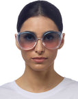A woman with dark hair parted in the middle wears oversized, round Okkia sunglasses in butterfly style with blue and pink gradient lenses. She has a neutral expression, wears a plain white shirt, and is set against a white background.