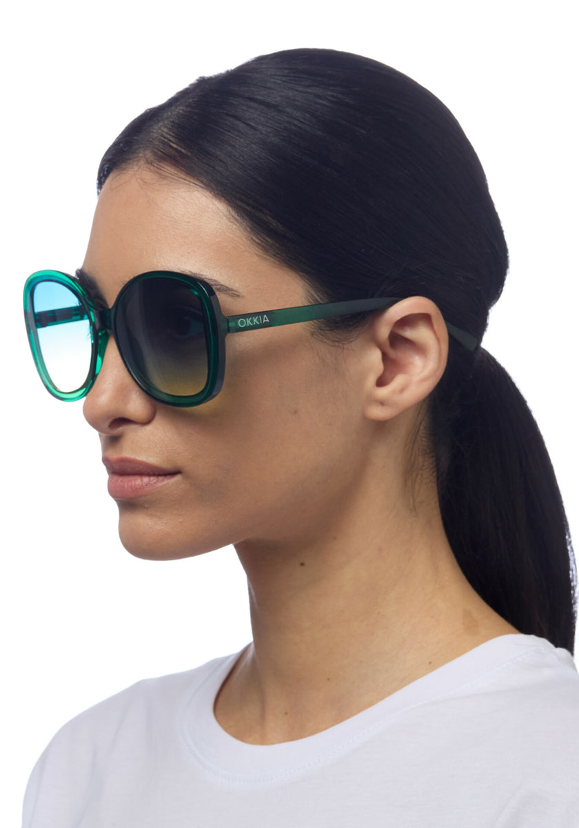A woman with straight dark hair tied back wears oversized Okkia grüne Butterfly-Sonnenbrille und ein schlichtes weißes Shirt. Seitlich vor weißem Hintergrund bieten die runden Sonnenbrillen stylischen UV-Schutz für ihre Augen.