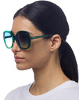 A woman with straight dark hair tied back wears oversized Okkia grüne Butterfly-Sonnenbrille und ein schlichtes weißes Shirt. Seitlich vor weißem Hintergrund bieten die runden Sonnenbrillen stylischen UV-Schutz für ihre Augen.