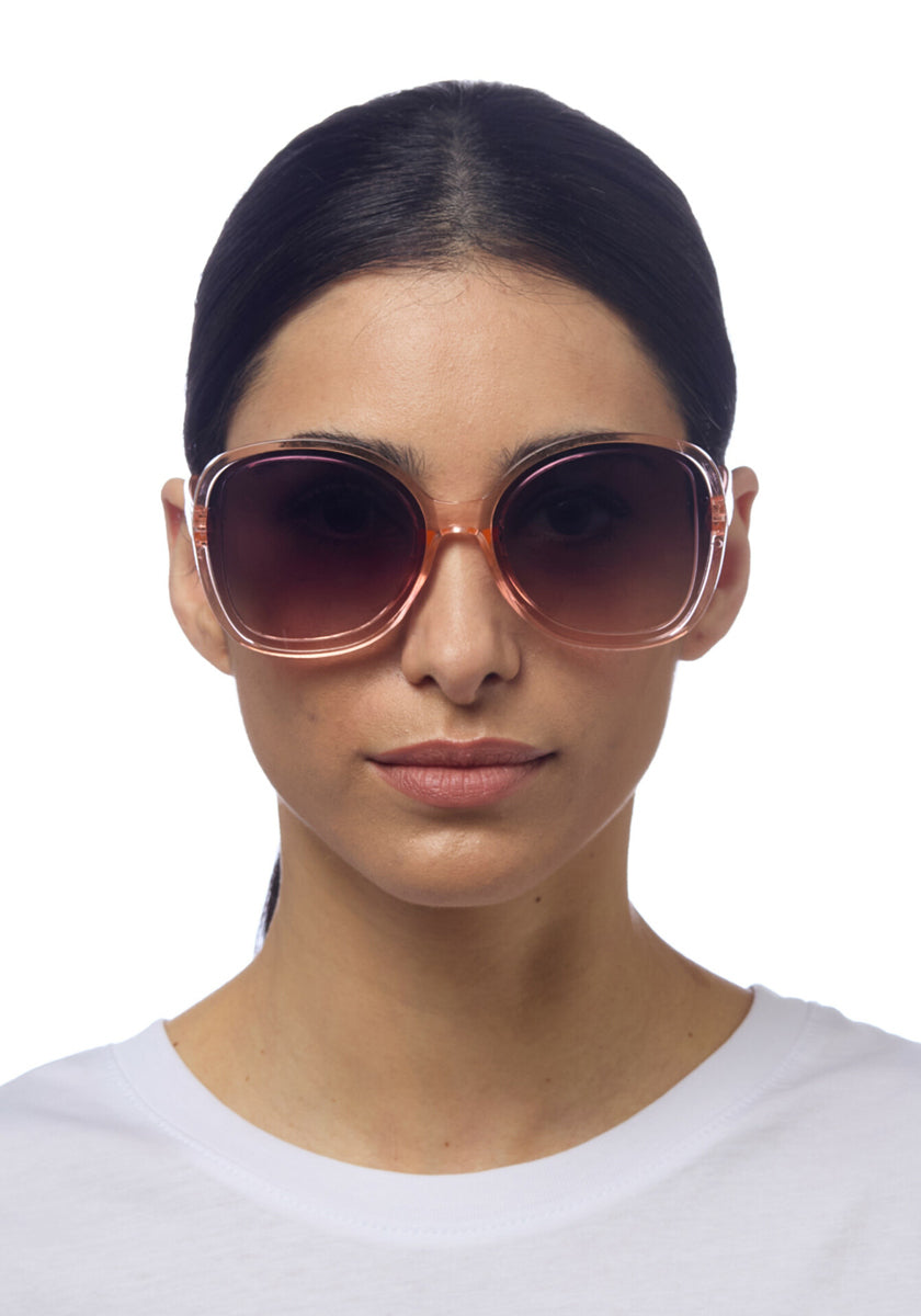 A woman with straight dark hair pulled back wears Okkia oversized round butterfly-style sunglasses with UV protection and a translucent frame. She has a neutral expression and is dressed in a plain white top against a white background.