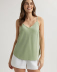 A woman with long brown hair smiles against a plain light background, wearing a light green Bella Dahl Lyocell Top with verstellbare Träger and white shorts. One hand rests by her side, the other on her thigh.
