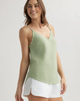 A woman with light skin and brown hair wears a light green Bella Dahl Lyocell Top with frayed edges and adjustable straps, paired with white shorts featuring curved side slits. She stands against a plain background, smiling and gazing left.
