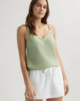 A woman with long brown hair wears a light green Bella Dahl Lyocell Top with lace trim and adjustable straps, paired with white drawstring shorts. She stands against a plain light background, looking at the camera with a relaxed expression.