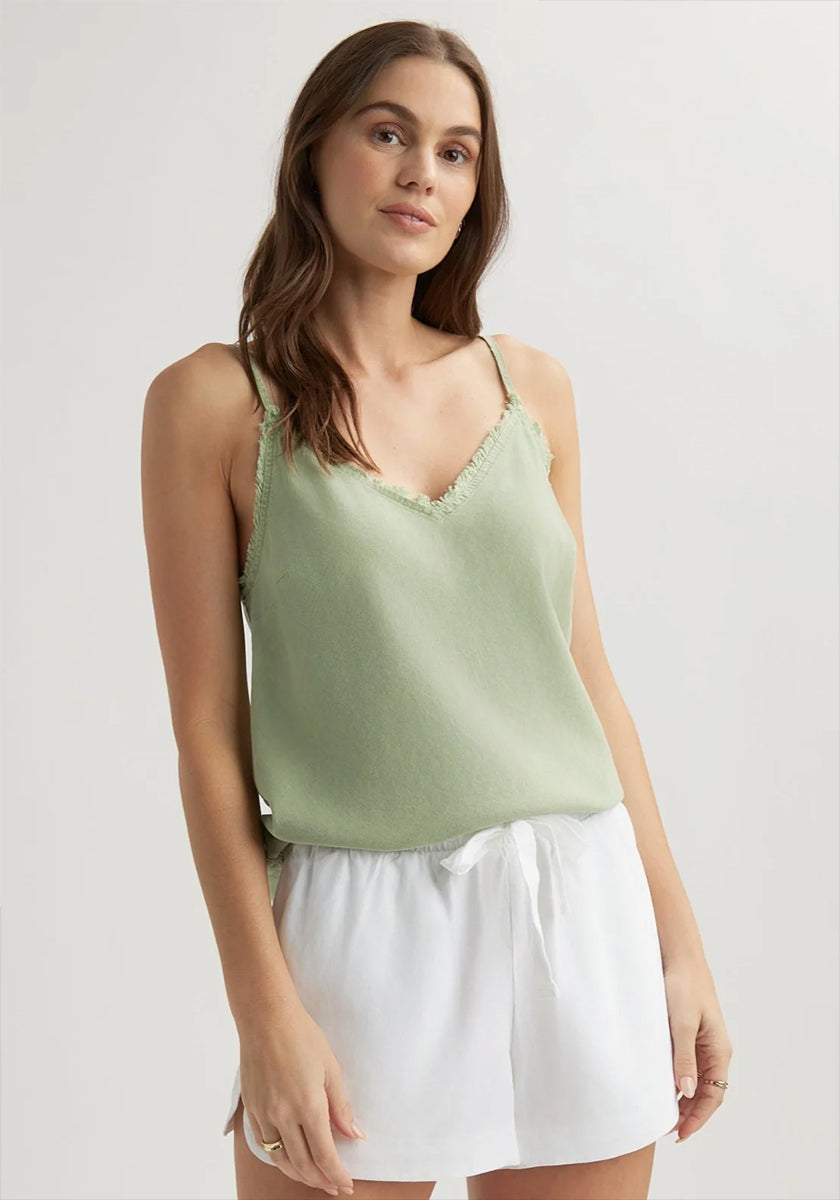 A woman with long brown hair and light skin stands against a plain background wearing a Bella Dahl light green Lyocell Trägertop with frayed edges and loose white drawstring shorts. She has a neutral expression and one hand by her side.