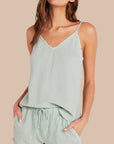 A woman models a relaxed-fit Bella Dahl Lyocell sage green Trägertop with thin straps and matching drawstring shorts in denim look, both featuring subtle frayed edges, as she poses with hands in pockets against a beige background.