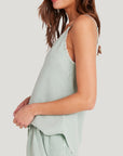 A woman with wavy brown hair, seen from the side, models a light sage green Bella Dahl Lyocell Trägertop and matching loose pants. The relaxed-fit top features raw-edge detailing on the armholes and neckline, set against a plain light gray background.