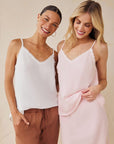 Two women stand side by side, smiling. One wears a white Bella Dahl Lyocell Trägertop with brown pants, her hair tied back. The other sports a light pink Bella Dahl camisole and skirt set, both tops featuring delicate straps and subtle lace trim.