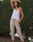 A smiling woman stands barefoot outdoors by a green hedge, wearing a white Bella Dahl Trägertop and loose beige Lyocell jogger pants, one hand in her pocket and the other on her head for a bright, casual look.