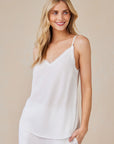 A woman with long blonde hair smiles in front of a light beige background, wearing a white Bella Dahl Lyocell Trägertop with a V-neckline and delicate trim, paired with matching white pants. Her hand rests by her side.