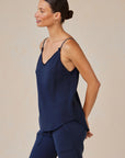 A woman with a low bun wears a sleeveless navy blue Lyocell V-neck camisole and matching pants from Bella Dahl. She stands in profile, looking left, arms relaxed behind her, against a light beige background.