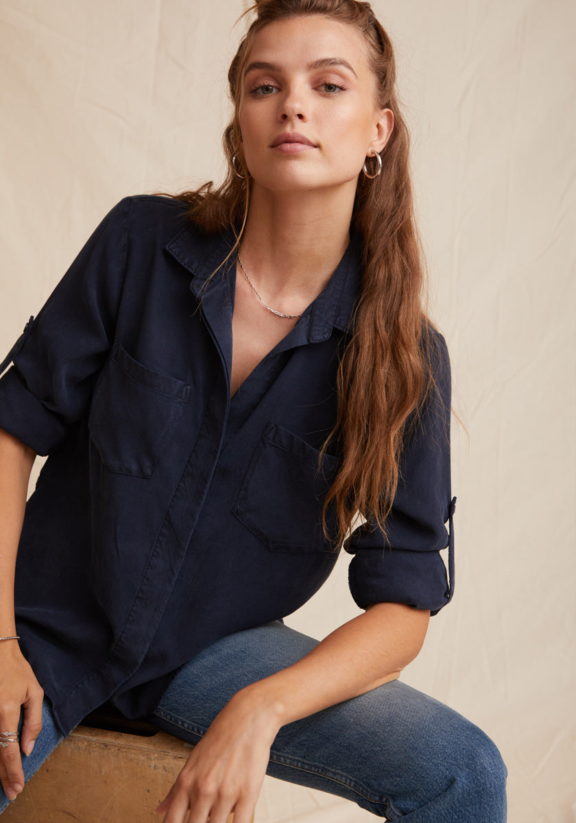 A woman with long, wavy light brown hair wears a navy Denimoptik Hemd aus 100% Lyocell by Bella Dahl, featuring rolled-up sleeves and Brusttaschen, paired with blue jeans. She sits on a stool against a beige background, looking confidently at the camera.