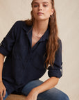 A woman with long, wavy light brown hair wears a navy Denimoptik Hemd aus 100% Lyocell by Bella Dahl, featuring rolled-up sleeves and Brusttaschen, paired with blue jeans. She sits on a stool against a beige background, looking confidently at the camera.