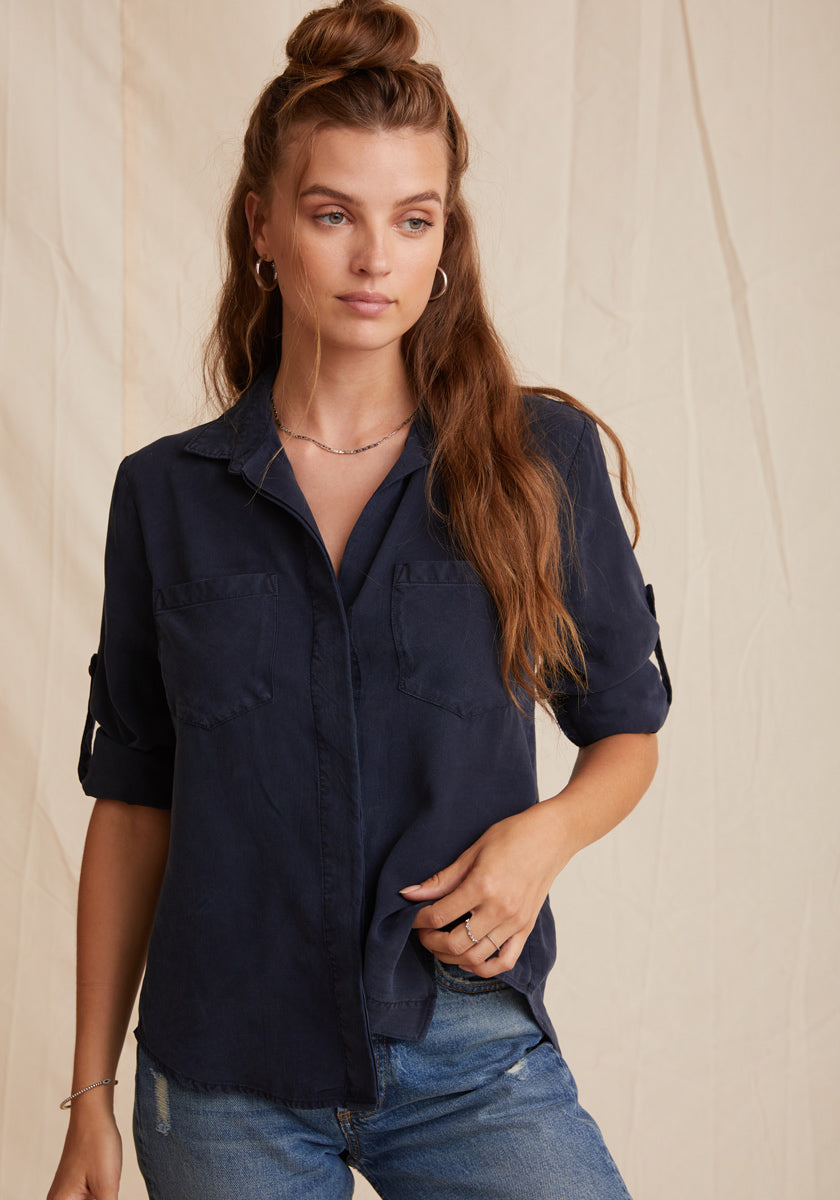 A woman with long, wavy brown hair wears a Bella Dahl navy Denimoptik Hemd with Brusttaschen and blue jeans. She stands against a light beige, slightly wrinkled fabric backdrop, gazing to her left with a relaxed expression.