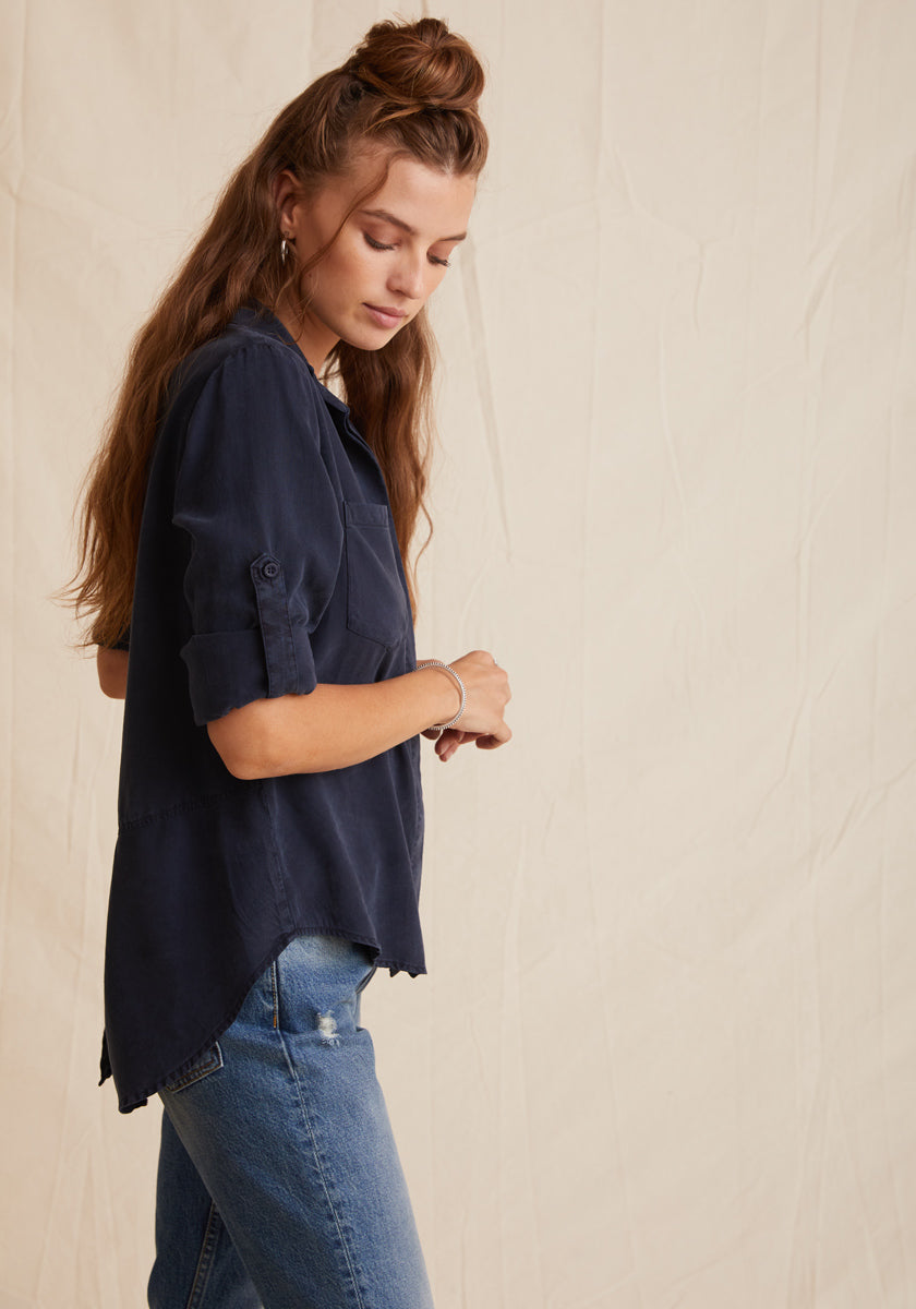 A woman with long, wavy brown hair models a navy Lyocell button-up shirt by Bella Dahl with a classic collar and rolled sleeves, paired with blue jeans. She stands sideways, looking down against a beige, slightly wrinkled backdrop.