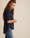 A woman with long, wavy brown hair models a navy Lyocell button-up shirt by Bella Dahl with a classic collar and rolled sleeves, paired with blue jeans. She stands sideways, looking down against a beige, slightly wrinkled backdrop.