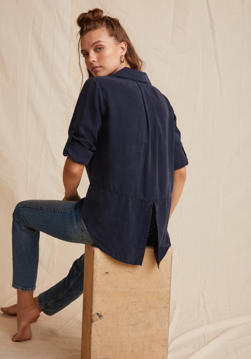 A light-skinned woman with long brown hair, half tied up, sits sideways on a wooden block against a beige draped background. She wears a navy blue Bella Dahl Lyocell button-up shirt with rolled sleeves and back slit, plus fitted jeans.