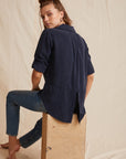 A light-skinned woman with long brown hair, half tied up, sits sideways on a wooden block against a beige draped background. She wears a navy blue Bella Dahl Lyocell button-up shirt with rolled sleeves and back slit, plus fitted jeans.