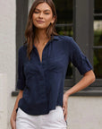 A woman with long brown hair stands outdoors in front of a white wall and black-trimmed window, wearing a navy blue Bella Dahl Denimoptik Hemd aus 100% Lyocell with rolled-up sleeves and Brusttaschen, paired with white pants.