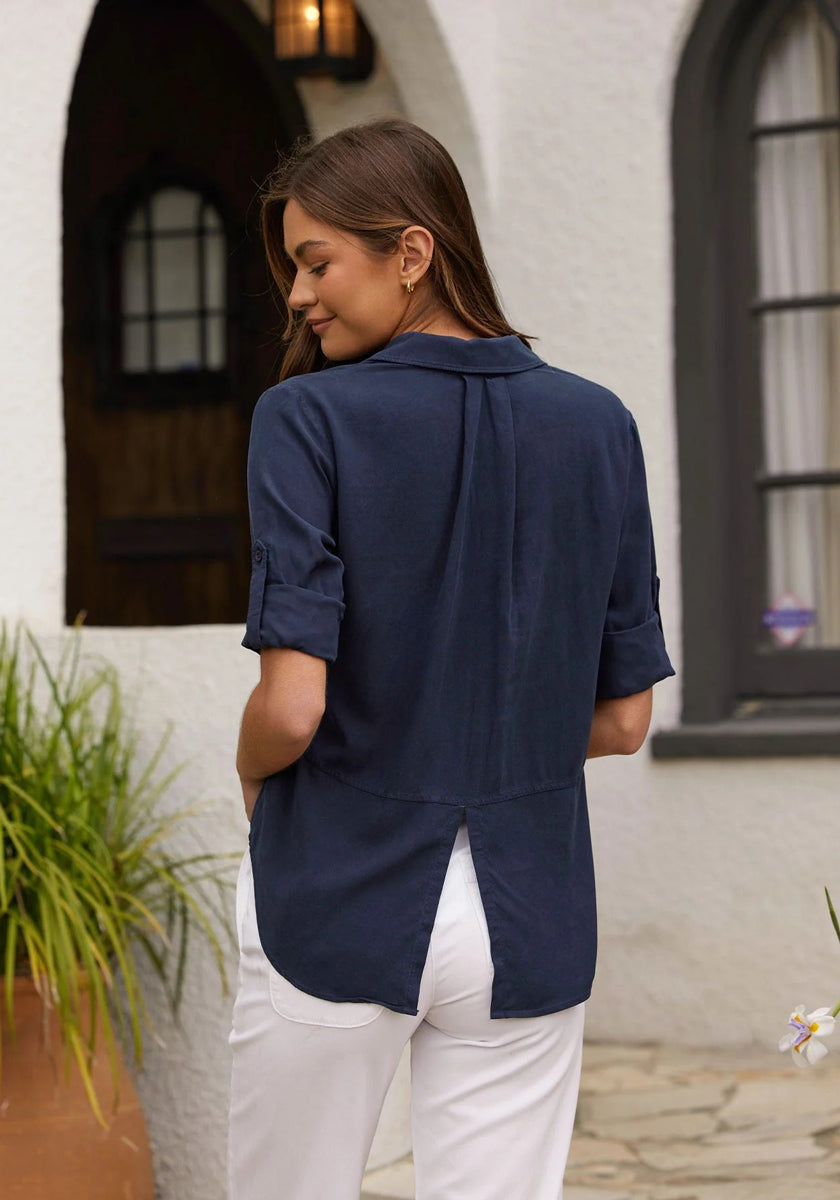 A woman with straight brown hair stands outdoors facing away, wearing a Bella Dahl 100% Lyocell Denimoptik Hemd with rolled sleeves and back pleat, paired with white pants. Behind her is a building with arched windows and green plants.