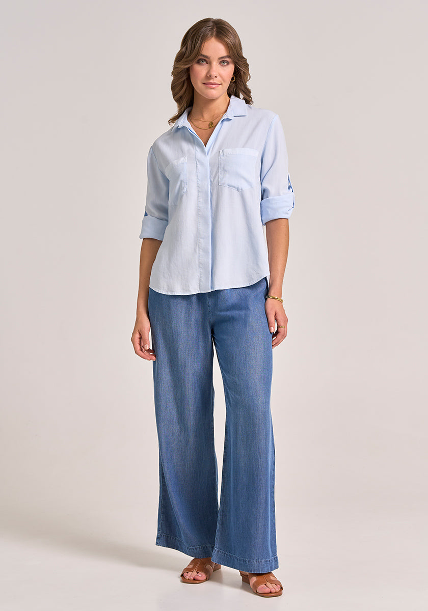 A woman with wavy brown hair stands against a plain background wearing a Bella Dahl light blue Lyocell button-up shirt with klassischer Hemdkragen and rolled sleeves, wide-leg Denimoptik jeans, brown sandals, plus a bracelet and necklace.