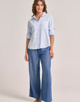 A woman with wavy brown hair stands against a plain background wearing a Bella Dahl light blue Lyocell button-up shirt with klassischer Hemdkragen and rolled sleeves, wide-leg Denimoptik jeans, brown sandals, plus a bracelet and necklace.