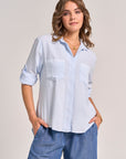 A woman with wavy light brown hair stands against a plain background, smiling gently as she wears gold jewelry and a Bella Dahl light blue button-up shirt with classic collar, two front pockets, and denim-look lyocell fabric.