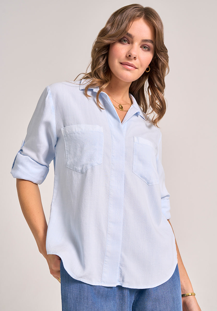 A woman with wavy light brown hair models a Bella Dahl light blue Denimoptik button-up shirt with a classic collar and rolled-up sleeves. She pairs it with blue jeans and gold jewelry, smiling gently against a plain background.