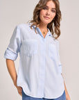 A woman with wavy light brown hair models a Bella Dahl light blue Denimoptik button-up shirt with a classic collar and rolled-up sleeves. She pairs it with blue jeans and gold jewelry, smiling gently against a plain background.