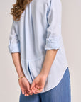 A person stands with their back to the camera in a Bella Dahl light blue Lyocell long-sleeve shirt with klassische Hemdkragen and rolled-up sleeves, paired with jeans. Their hands are clasped behind their back against a plain background.