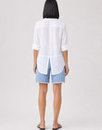 Eine Person steht mit dem Rücken zur Kamera und trägt ein weißes Bella Dahl Damenhemd mit umgeschlagenen Ärmelbündchen, blaue Jeansshorts mit ausgefransten Säumen und braune Sandalen. Sie hat glattes, schwarzes, schulterlanges Haar und posiert vor einem schlichten hellgrauen Hintergrund.